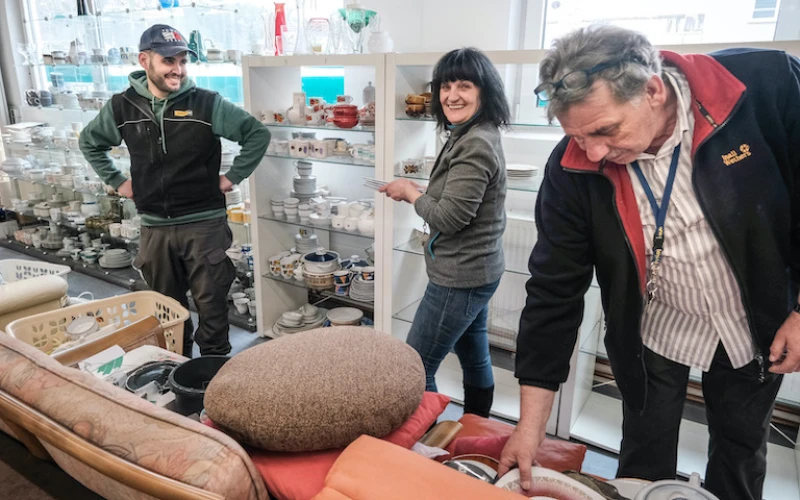 Drei Personen stehen in einem Sozialkaufhaus vor Regalen mit Geschirr. Die Personen scheinen bei der Arbeit oder Inventur zu sein.