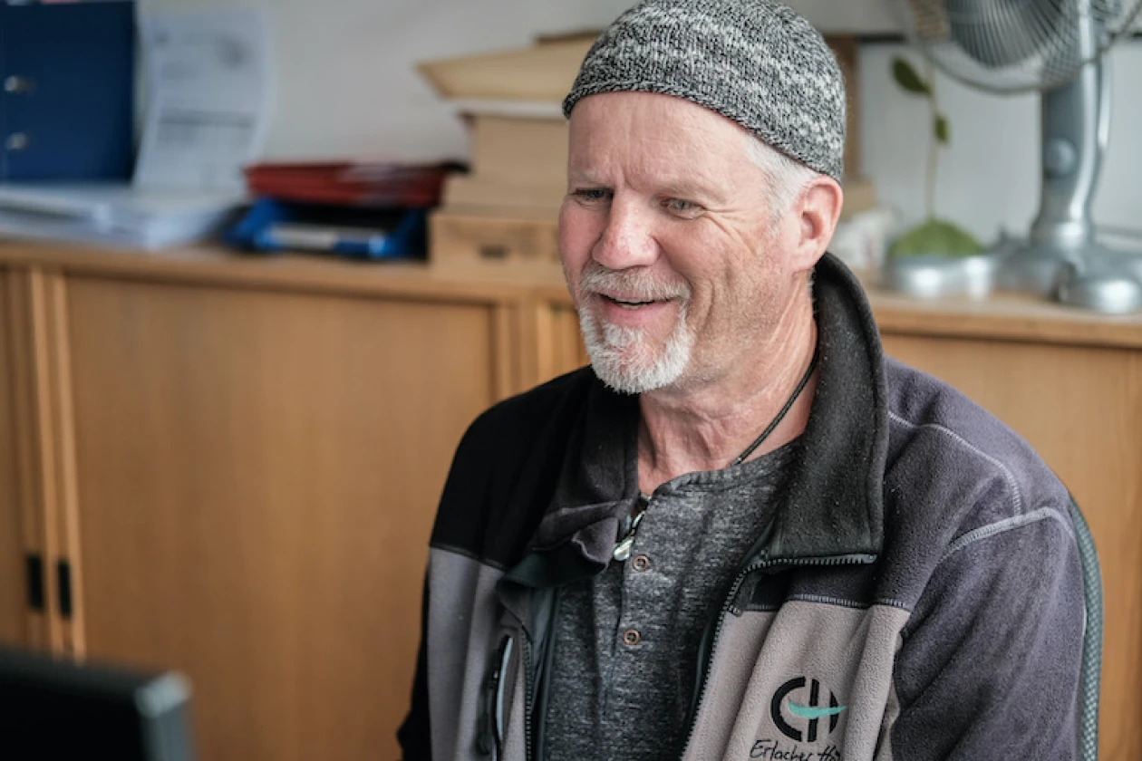 Im Vordergrund ist ein freundlich l&auml;chelnder Mitarbeiter der Erlacher H&ouml;he zu sehen. Er tr&auml;gt eine graue Strickm&uuml;tze und eine dunkele Jacke mit dem Erlacher H&ouml;he Logo. Im Hintergrund sind ein Schrank und Arbeitsunterlagen zu sehen.