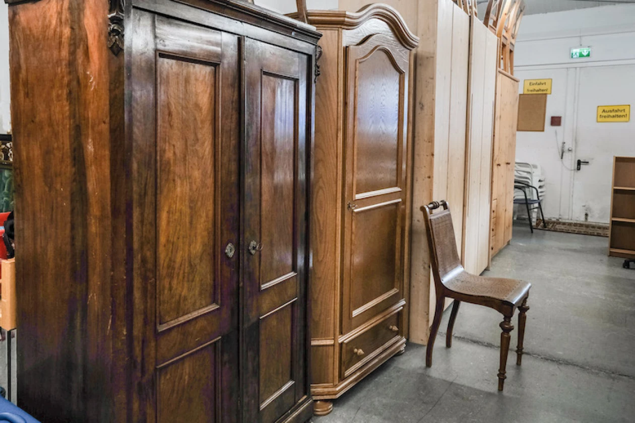 Massivholzschr&auml;nke in unterschiedlichen Braunt&ouml;nen stehen in einem Lagerraum. Ein einzelner Holzstuhl steht vor dem Schrank und im Hintergrund sind weitere Regale und ein Notausgangsschild &uuml;ber der T&uuml;r zu sehen.