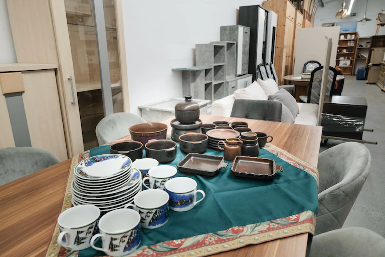 Tisch mit Keramikgeschirr und Tassen in verschiedenen Farben und Formen, im Hintergrund sind weitere verschiedene M&ouml;belst&uuml;cke zu sehen.