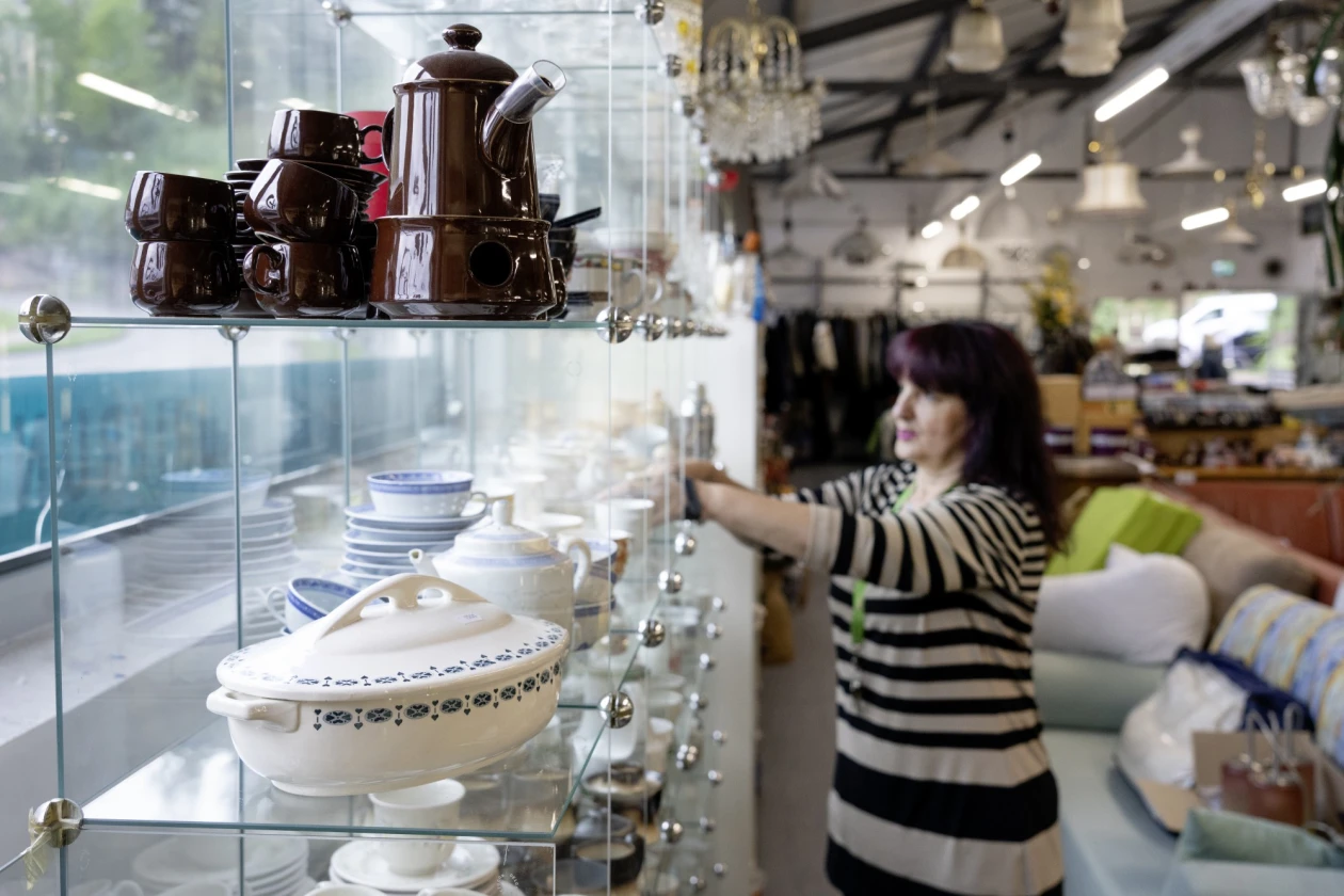 Wei&szlig;es Keramikgeschirr und eine braune Kaffeekanne auf Glasregalen in einem Gesch&auml;ft. Im Hintergrund eine Person in einem gestreiften Oberteil, die in einem unscharfen Regalbereich steht.