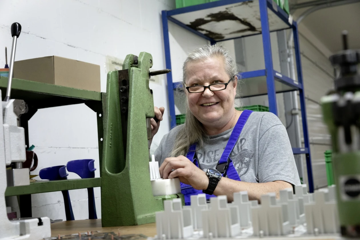 Eine &auml;ltere Frau mit Brille und grauem T-Shirt l&auml;chelt in einer Werkstatt an einer gr&uuml;nen Maschine. Sie tr&auml;gt eine blaue Arbeitshose und scheint zufrieden mit ihrer Arbeit zu sein. Die Umgebung ist eine professionelle Werkstatteinrichtung mit verschiedenen Maschinen und Regalen im Hintergrund.