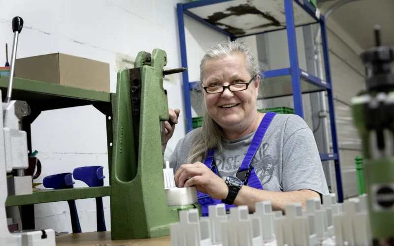 Eine &auml;ltere Frau mit Brille und grauem T-Shirt l&auml;chelt in einer Werkstatt an einer gr&uuml;nen Maschine. Sie tr&auml;gt eine blaue Arbeitshose und scheint zufrieden mit ihrer Arbeit zu sein. Die Umgebung ist eine professionelle Werkstatteinrichtung mit verschiedenen Maschinen und Regalen im Hintergrund.