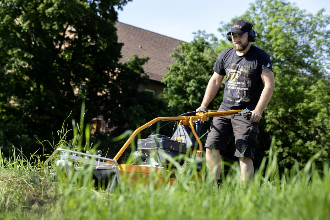 Ein Mann in Arbeitskleidung m&auml;ht Gras mit einem gelben Rasenm&auml;her an einem sonnigen Tag.