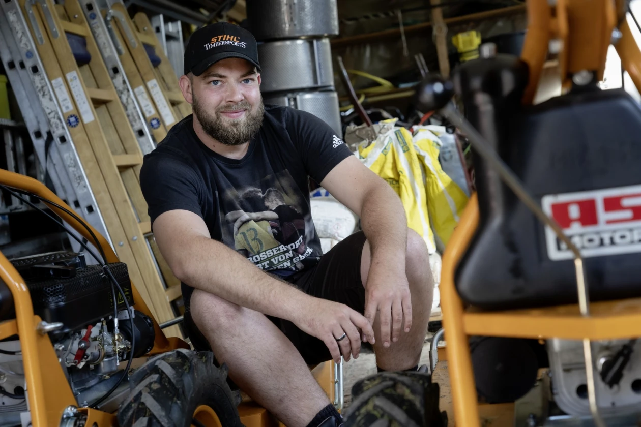 B&auml;rtiger Mann in schwarzem T-Shirt und Kappe sitzt in einem Raum zwischen Leitern und Bauger&auml;ten.