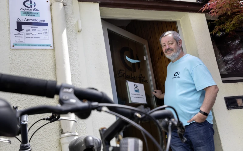 Ein Mann in einem T-Shirt mit Erlacher H&ouml;he Logo steht l&auml;chelnd an der Eingangst&uuml;r eines Geb&auml;udes, im Vordergrund ist ein Fahrradlenker zu sehen, daneben h&auml;ngt ein Schild von der Erlacher H&ouml;he mit dem Aufschrieb: "Ambulante Hilfen Rems-Murr".