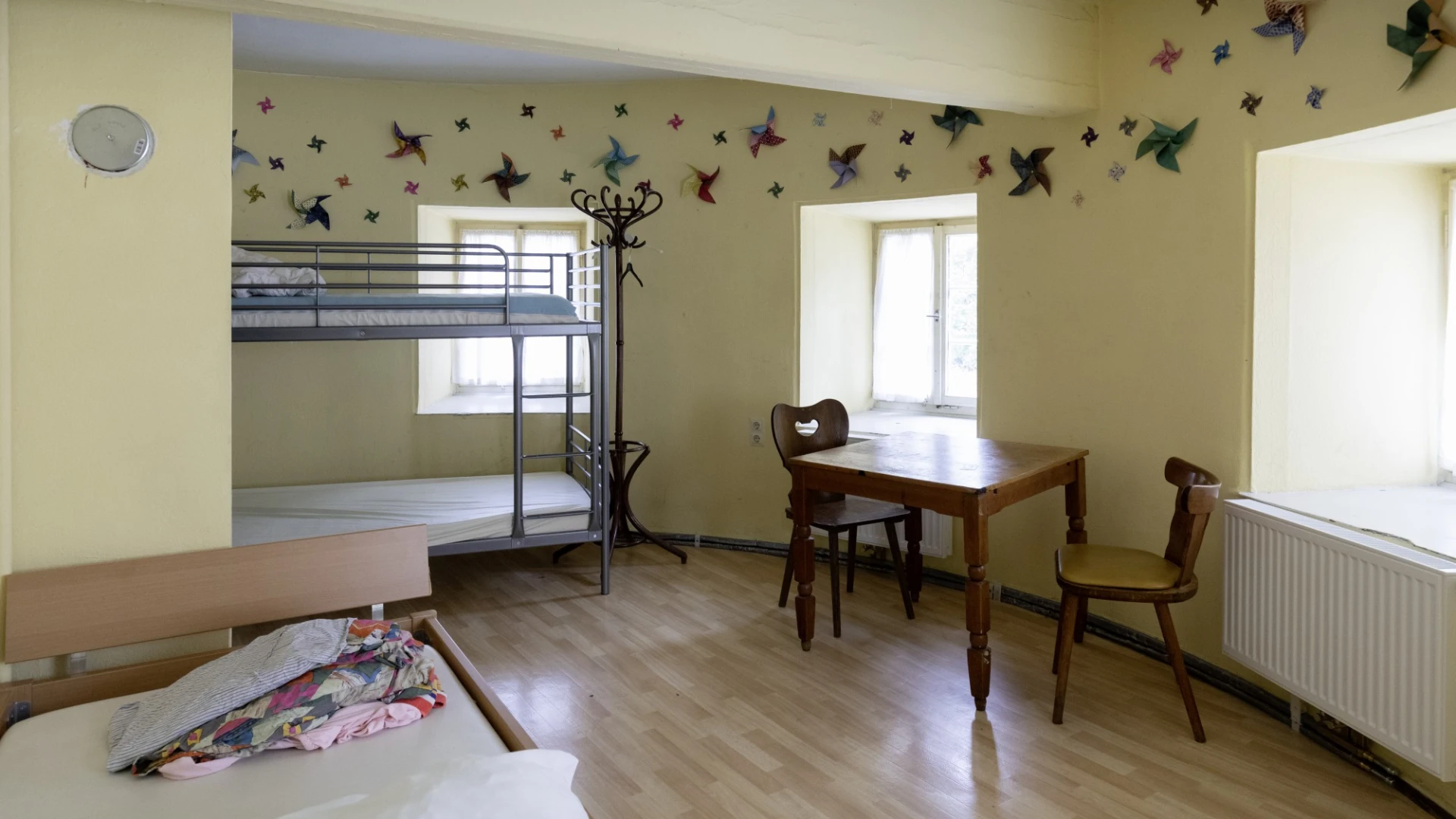 Zimmer mit Etagenbett, Einzelbett und einem kleinen Holztisch mit zwei St&uuml;hlen. Die W&auml;nde sind mit gelber Farbe gestrichen und mit bunten Schmetterlingen dekoriert.