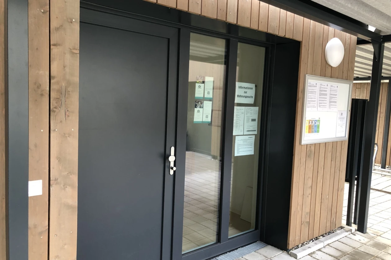 Moderner Eingangsbereich mit Holzfassade, schwarzer T&uuml;r mit Glasfenstern und einem &uuml;berdachten Vorbau. Ein Informationsschild h&auml;ngt rechts neben der Eingangst&uuml;r.
