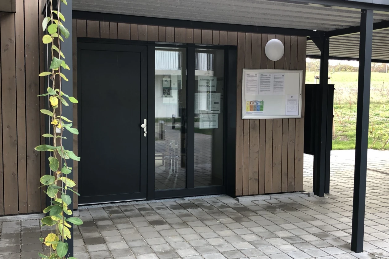 Moderner Eingangsbereich mit Holzfassade, schwarzer T&uuml;r mit Glasfenstern und einem &uuml;berdachten Vorbau. Links w&auml;chst eine Kletterpflanze mit gelben und gr&uuml;nen Bl&uuml;ten an der Wand. Ein Informationsschild h&auml;ngt rechts neben der Eingangst&uuml;r.