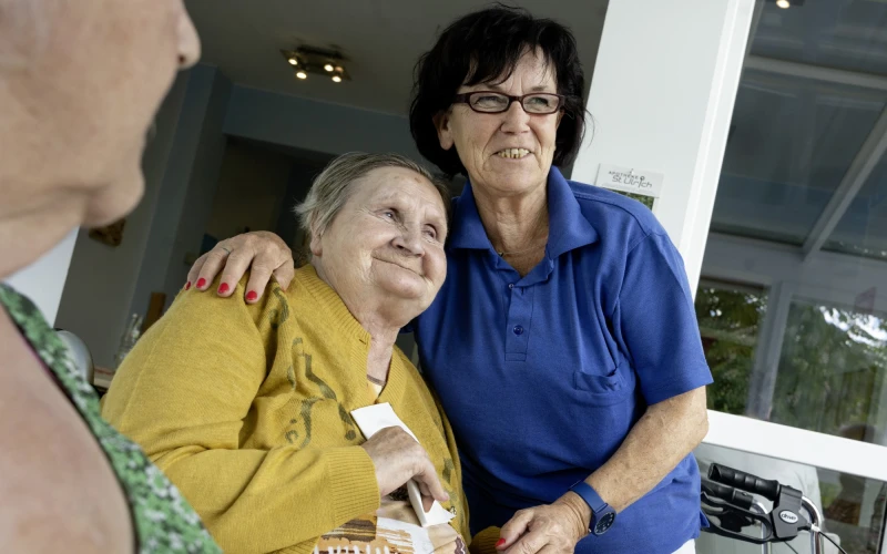 Zwei Frauen stehen eng beieinander und l&auml;cheln. Die eine tr&auml;gt ein gelbes, die andere ein blaues Polo-Shirt, sie legt den Arm um die andere Frau. Im Hintergrund sind ein Rollator und ein Fenster zu sehen.