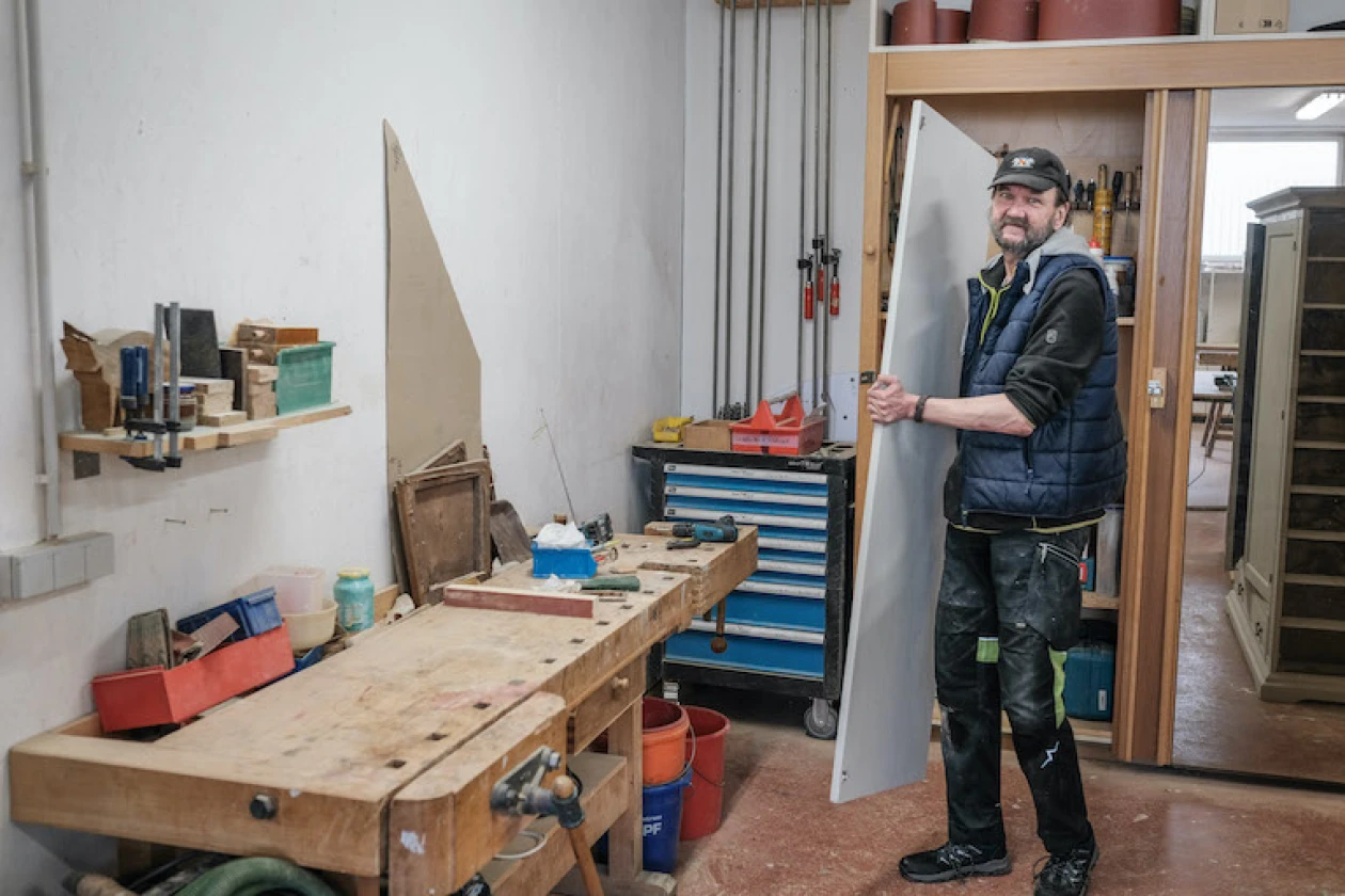 Ein Mann in Arbeitskleidung steht in einer Werkstatt neben einer Holzwerkbank umgeben von Werkzeugen, Regalen und einer blauen Werkzeugkiste. Er tr&auml;gt eine Weste und eine Kappe und h&auml;lt eine gro&szlig;e Holzplatte.