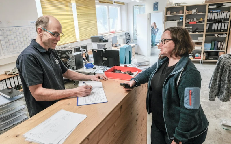 Ein Mitarbeiter steht hinter einem Holztresen und hat einen Kugelschreiber und ein Dokument in der Hand. Vor ihm steht eine Frau mit einem Telefon in der Hand. Beide tragen Brillen und arbeiten in einem hellen B&uuml;ro mit Regalen und Aktenordnern im Hintergrund.
