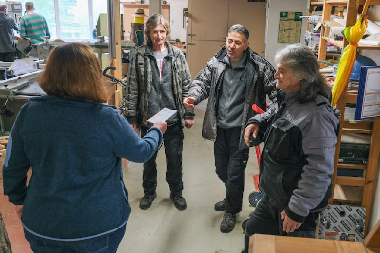 Vier Personen in Winterjacken stehen in einem Werkstattraum. Eine Frau in blauer Dienstkleidung reicht einer anderen Person ein Papier. Der Raum ist mit Regalen und Schr&auml;nken ausgestattet.