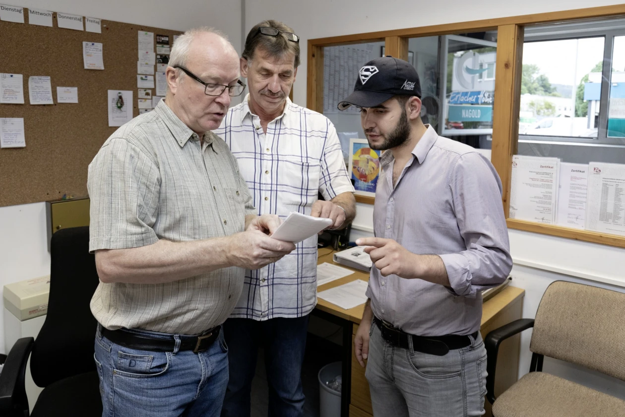 Drei M&auml;nner stehen in einem B&uuml;ro und betrachten gemeinsam ein Dokument. Im Hintergrund sind eine Pinnwand und ein Schreibtisch zu sehen.