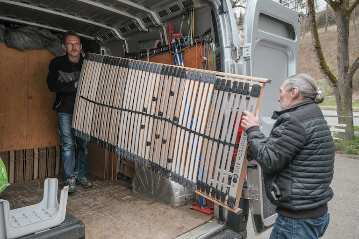 Eine Person steht auf der Ladefl&auml;che eines Transporters und h&auml;lt gemeinsam mit einer weiteren Person, die vor der ge&ouml;ffneten T&uuml;r steht, einen Lattenrost fest.