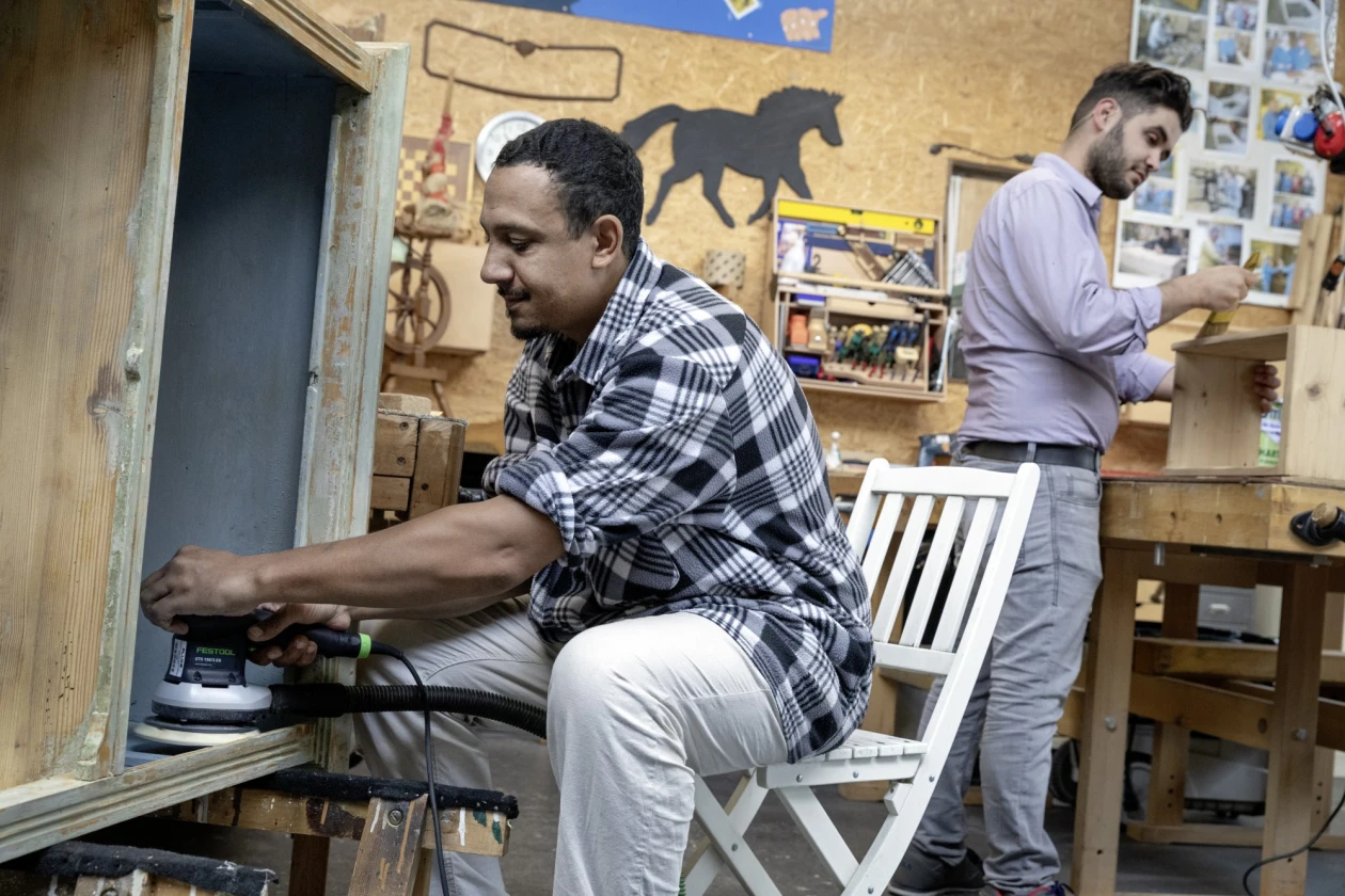 Zwei M&auml;nner arbeiten in einer Werkstatt. Ein Mann in kariertem Hemd sitzt auf einem wei&szlig;en Stuhl und arbeitet mit einer Schleifmaschine an einem Schrank, w&auml;hrend der andere Mann im Hintergrund mit einem Pinsel ein Regal streicht.