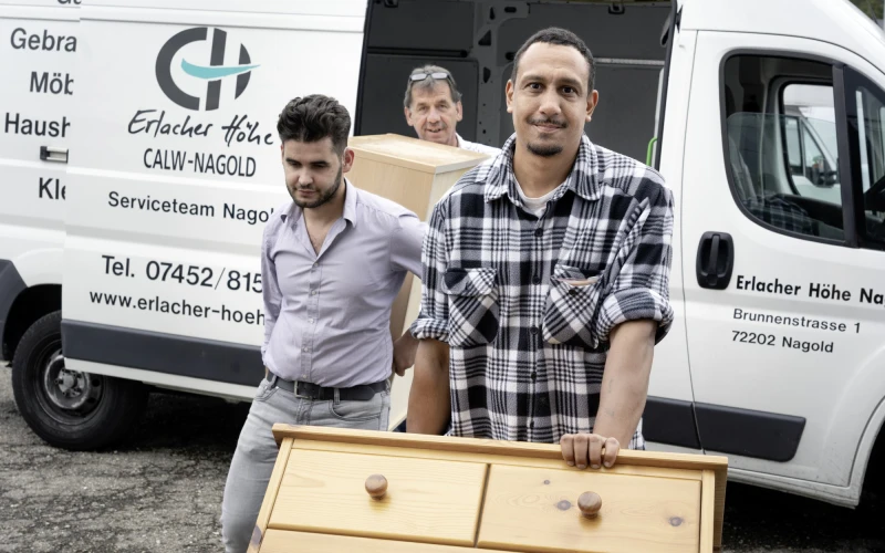 Drei Mitarbeiter des Serviceteams Nagold stehen vor einem Firmenfahrzeug. Der Mann vorn hat seine Hand auf einem Schrank, w&auml;hrend die beiden hinter ihm gemeinsam ein M&ouml;belst&uuml;ck tragen.