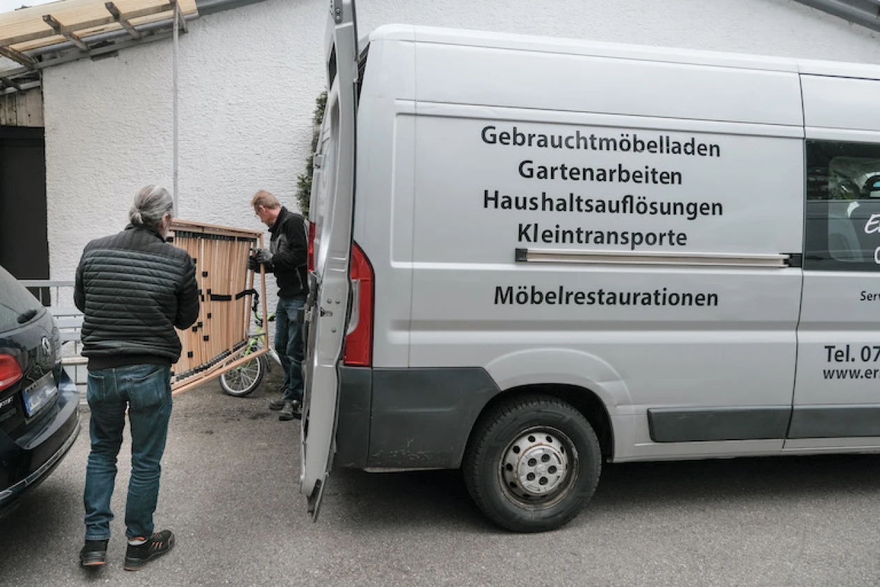 Zwei M&auml;nner stehen neben einem wei&szlig;en Transporter mit verschiedenen Dienstleistungsaufschriften wie Gartenarbeiten und M&ouml;beltransporte. Der Transporter ist auf einem Parkplatz geparkt, und die zwei M&auml;nner tragen gemeinsam einen Lattenrost.