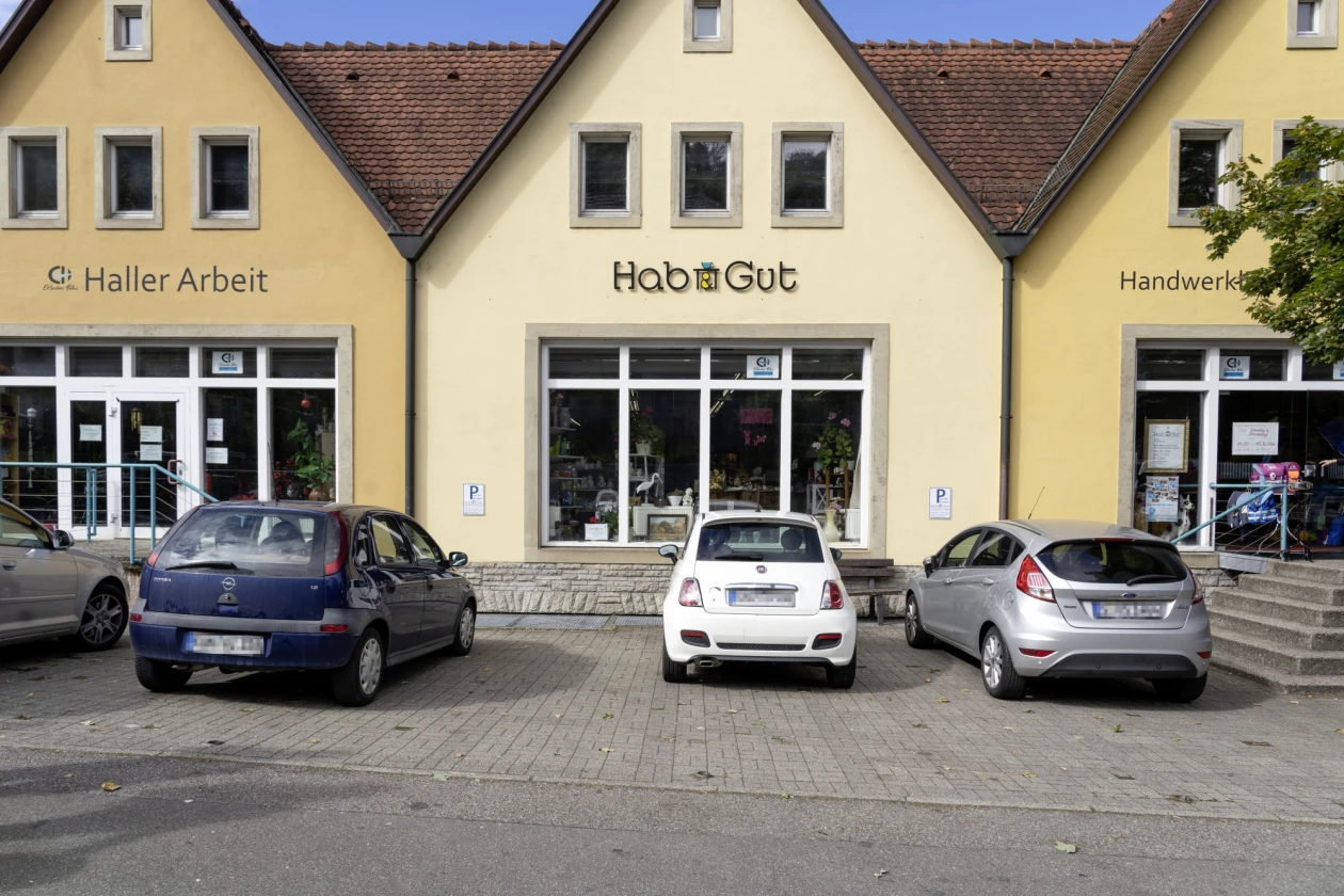 Vier Autos parken vor gelben Gesch&auml;ftsgeb&auml;uden der Erlacher H&ouml;he mit verschiedenen Ladenfronten wie "Haller Arbeit" und "Hab und Gut". Die Fassaden sind einheitlich gestaltet mit grauen Fenstern.