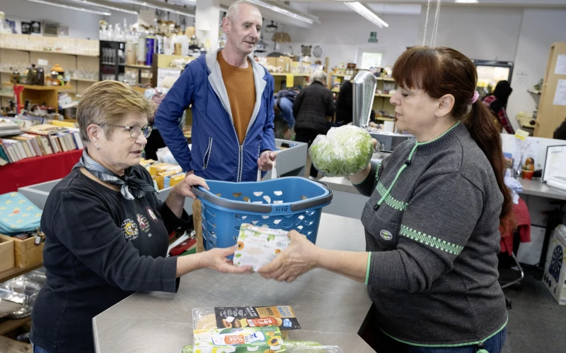 Im Vordergrund sind zwei Frauen an einer Ladentheke zu sehen. Die eine Frau h&auml;lt einen verpackten Salat in der Hand und legt ihn in den blauen Einkaufskorb der anderen Frau.