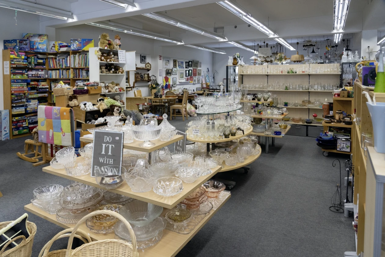 Ein Raum in einem Sozialkaufhaus mit Holzregalen und Tischen, auf denen verschiedene Keramik-, Glas- und Kunsthandwerksobjekte ausgestellt sind. Regale im Hintergrund sind mit B&uuml;chern gef&uuml;llt.