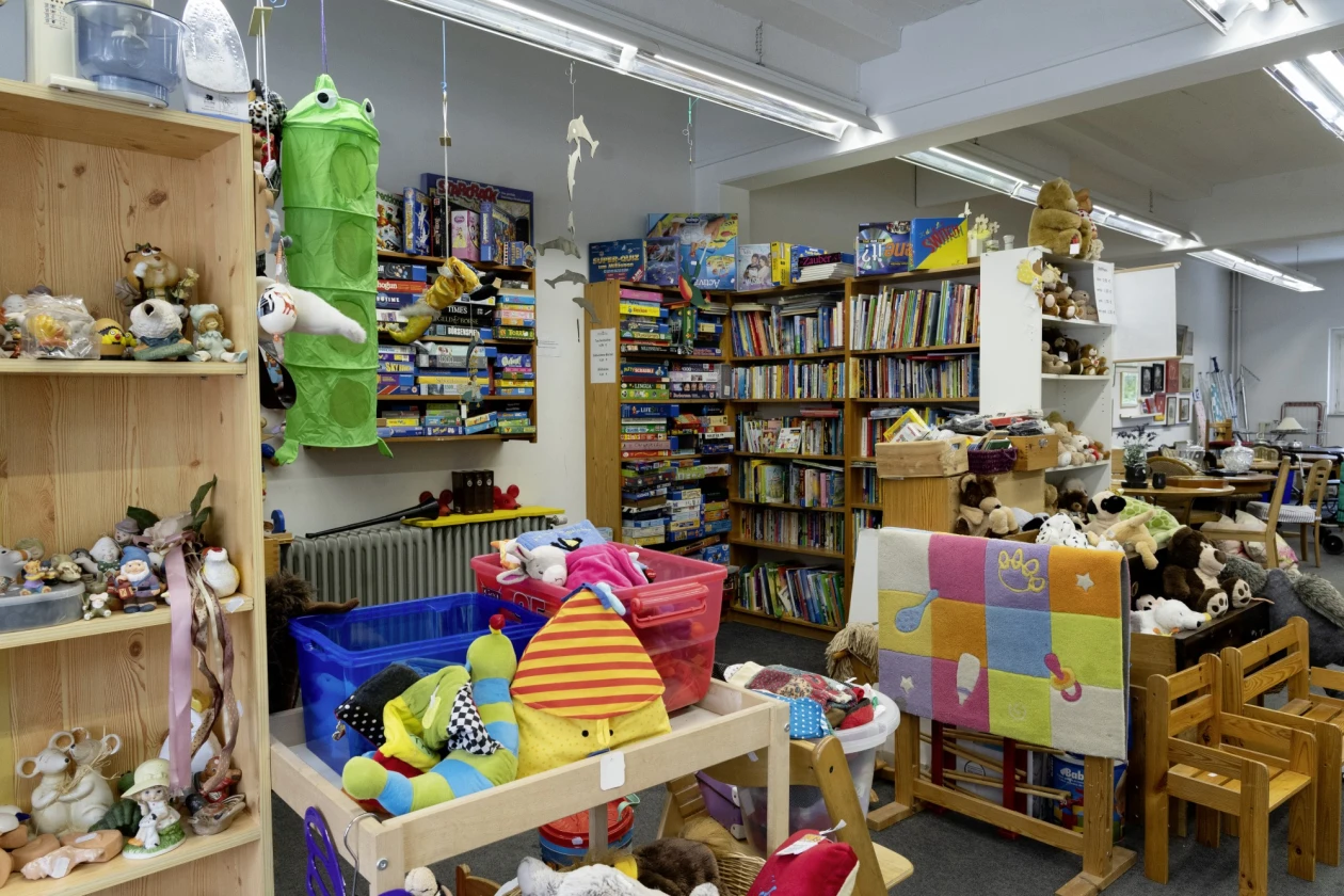 Ein Raum in einem Sozialkaufhaus mit vielen Spielzeugen, B&uuml;cherregalen und bunten Aufbewahrungsk&ouml;rben. Verschiedene Pl&uuml;schtiere und Stofftiere sind auf Regalen und Tischen verteilt.