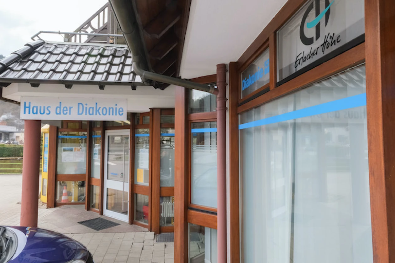 Eingang zum Sozialkaufhaus mit Glasfassade, davor ein Gehweg und Teil eines parkenden Fahrzeugs.