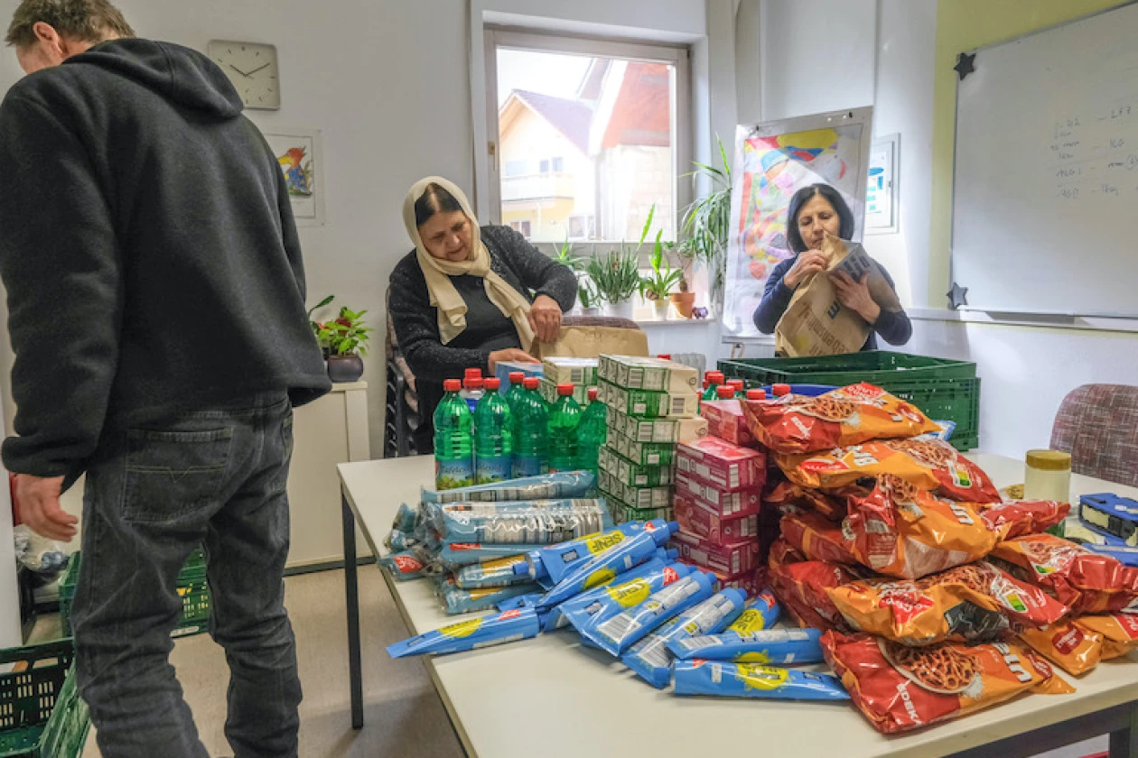An einem Tisch in einem hellen Raum stehen 3 Personen. Auf dem Tisch stehen Lebensmittel und Getr&auml;nke. Zu sehen sind unter anderem gr&uuml;ne Flaschen, Tuben, T&uuml;ten und weitere verpackte Produkte.