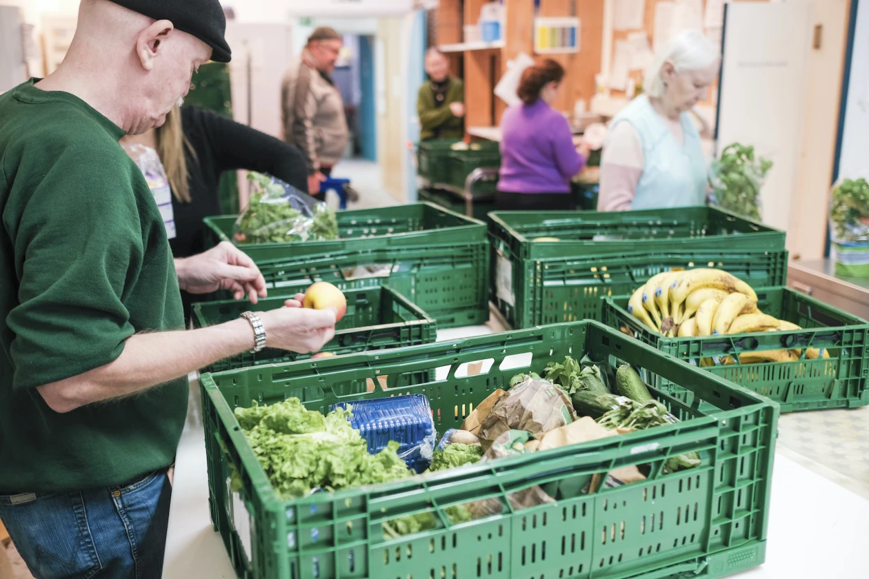 Auf einem Tisch stehen gr&uuml;ne Plastikkisten mit Gem&uuml;se und Obst. Ein Mann in gr&uuml;nem Pullover h&auml;lt einen Apfel in seiner rechten Hand. Im Hintergrund sind weitere Personen zu sehen.