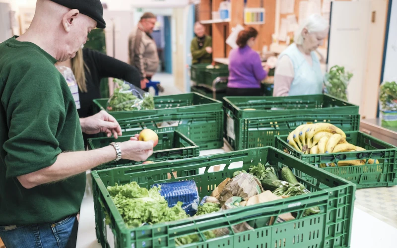 Auf einem Tisch stehen gr&uuml;ne Plastikkisten mit Gem&uuml;se und Obst. Ein Mann in gr&uuml;nem Pullover h&auml;lt einen Apfel in seiner rechten Hand. Im Hintergrund sind weitere Personen zu sehen.