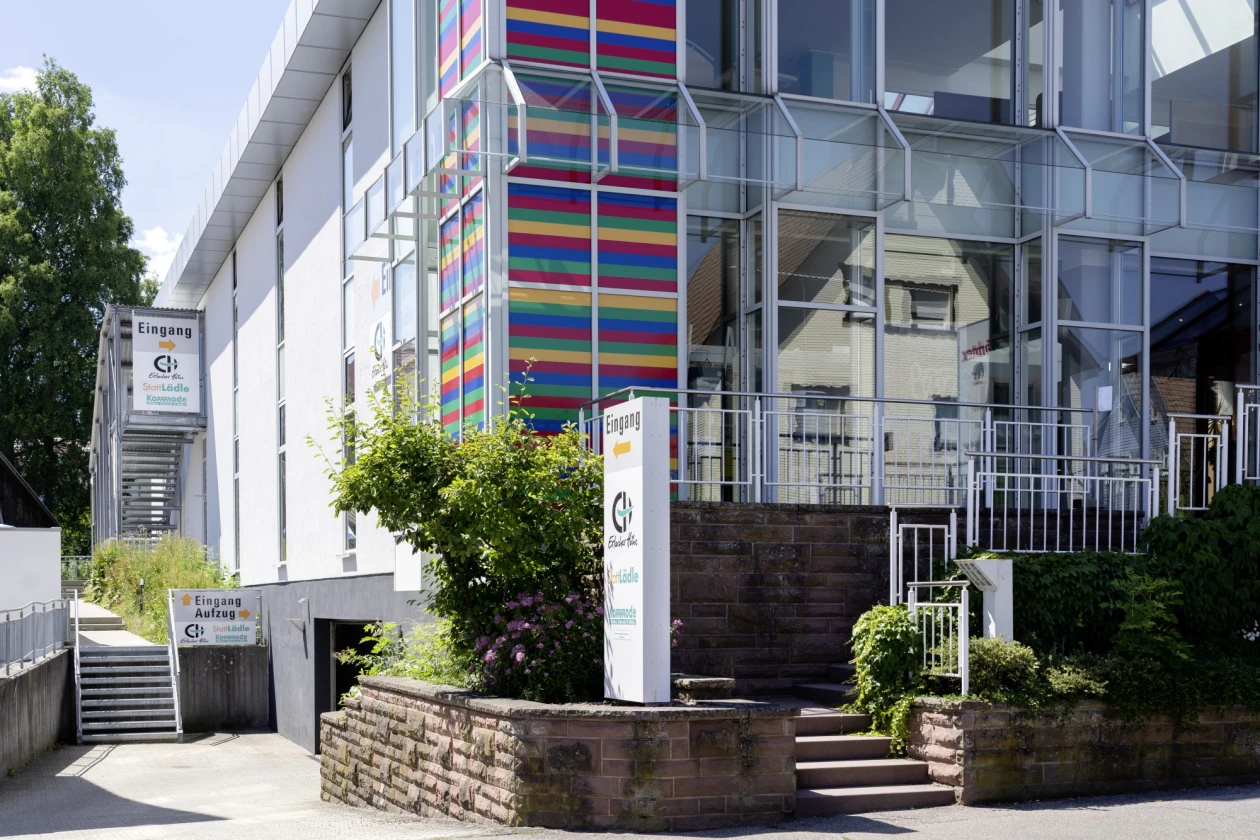 Ein Geb&auml;ude der Erlacher H&ouml;he, mit einem bunten Fassadenstreifen, umgeben von Gr&uuml;npflanzen und Steinmauern. Eine Treppe mit Gel&auml;ndern f&uuml;hrt zum Eingangsbereich.