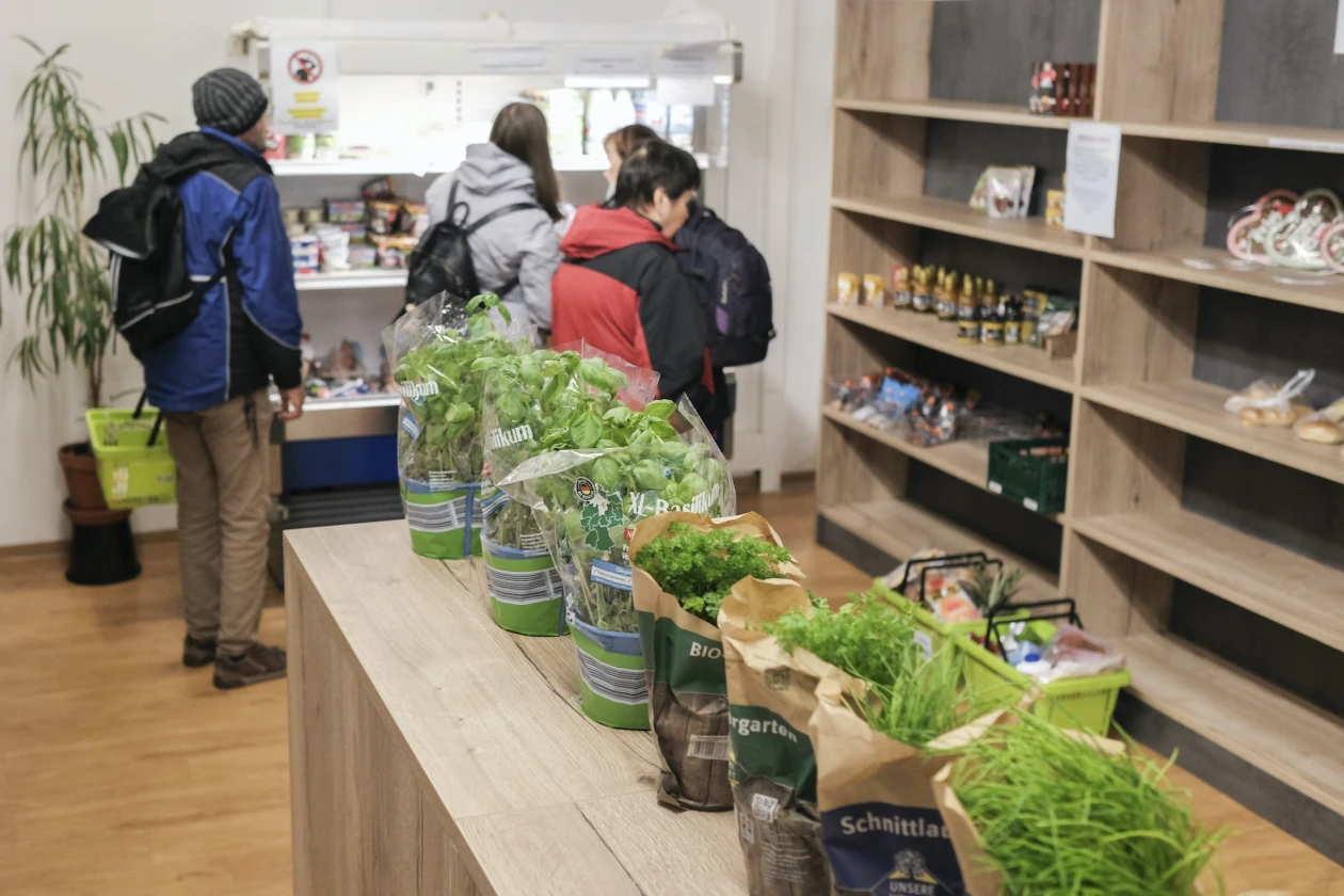 Mehrere Personen stehen an einem K&uuml;hlregel. Im Vordergrund stehen T&ouml;pfe mit frischen Kr&auml;utern auf einer Theke. Dahinter stehen Holzregale, in denen verpackte Lebensmittel pr&auml;sentiert werden.