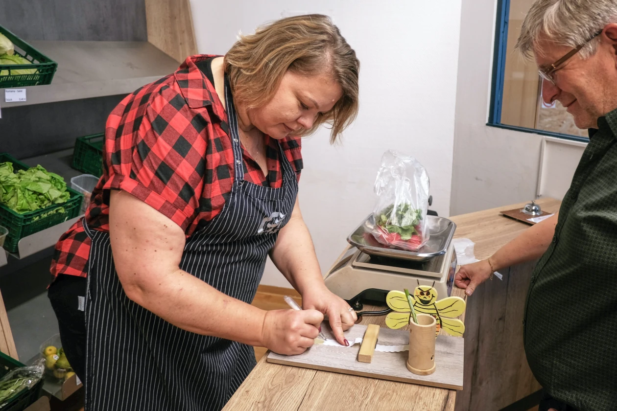 Vor einem Gem&uuml;seregal steht eine Frau in kariertem Hemd und gestreifter Sch&uuml;rze an einer Theke, h&auml;lt einen Kugelschreiber in der Hand und schreibt ein Preisetikett f&uuml;r abgewogenes Gem&uuml;se. Ein Mann steht gegen&uuml;ber und beobachtet sie.