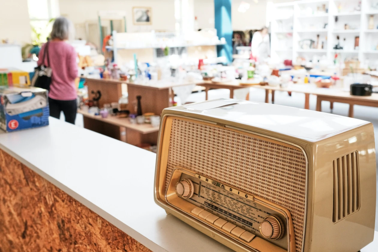 Zu sehen ist ein Raum mit verschiedenen Regalen und Tischen voller Materialien. Im Vordergrund steht ein altes Radio auf einer Holztheke mit wei&szlig;er Oberfl&auml;che.