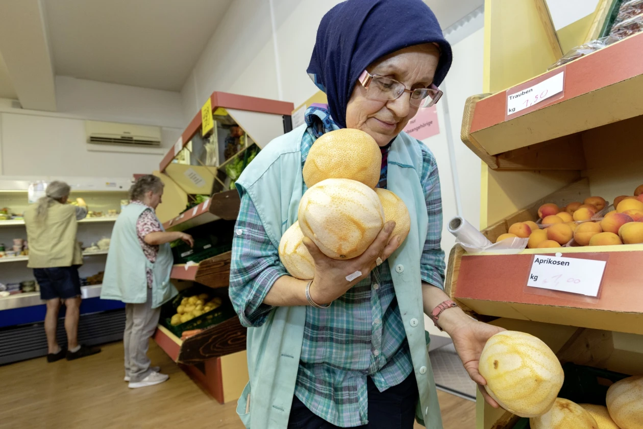 Eine &auml;ltere Frau mit Kopftuch und Brille h&auml;lt in einem Laden mehrere gelbe Obstst&uuml;cke in den Armen. Im Hintergrund befinden sich weitere Regale mit Obst und Gem&uuml;se sowie zwei weitere Personen.