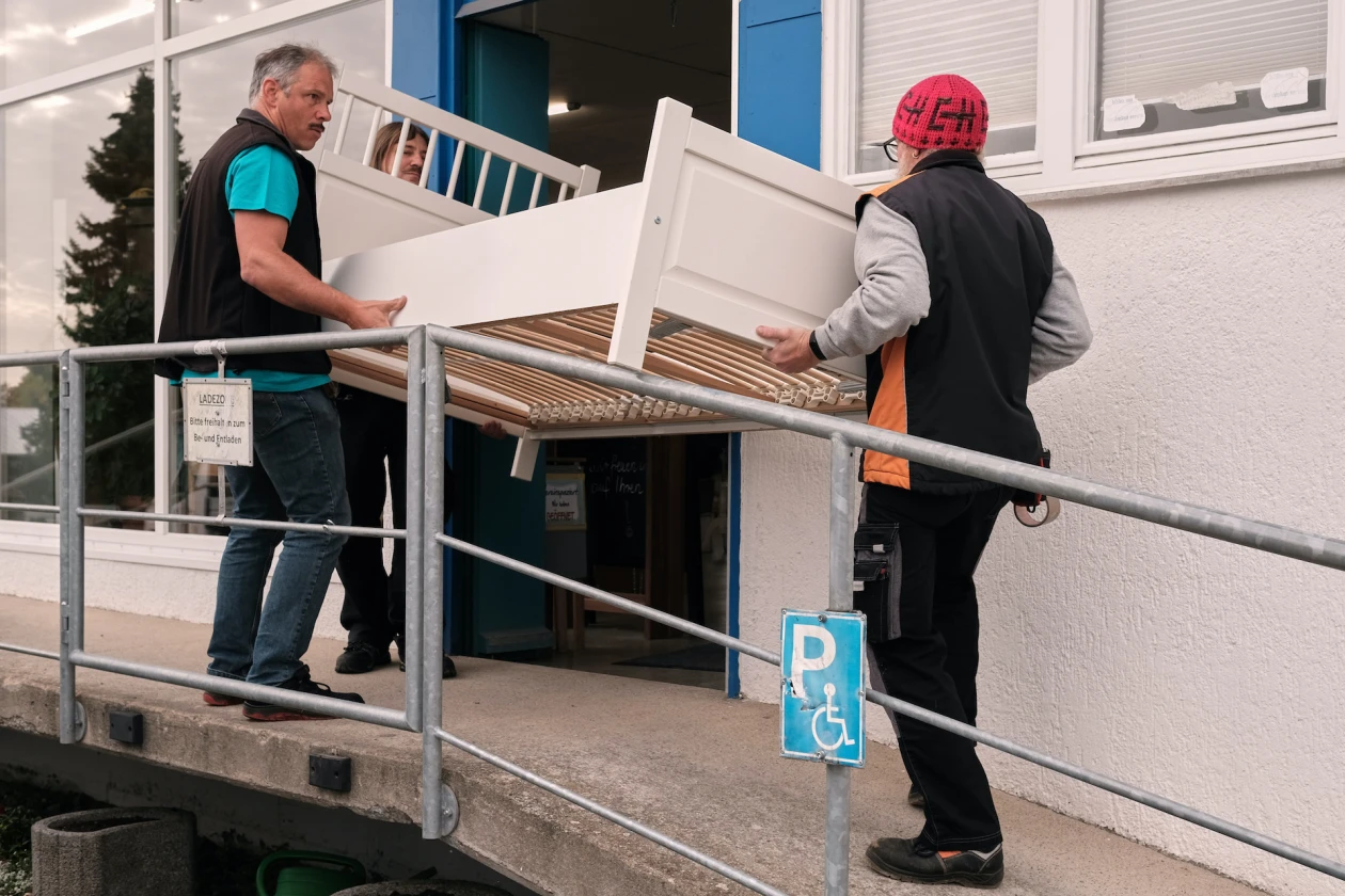Drei Personen tragen gemeinsam ein wei&szlig;es Bett. Sie stehen vor der Eingangst&uuml;r zu einem Geb&auml;ude.