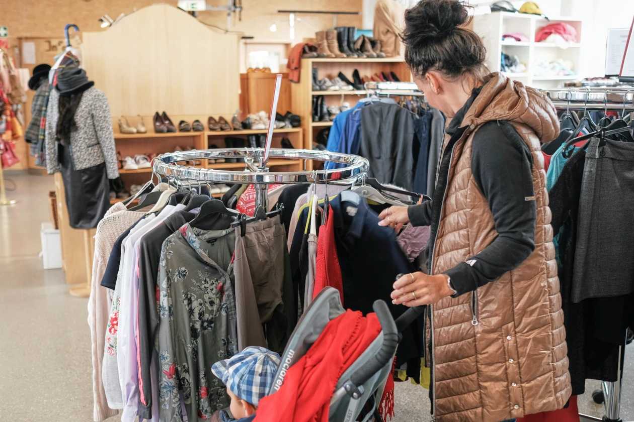 Eine Frau mit einem Kind in einem Kinderwagen schaut sich die Kleidung auf einem Kleiderst&auml;nder an. Im Hintergrund sind weitere Kleiderst&auml;nder sowie Regale mit Schuhen zu sehen.