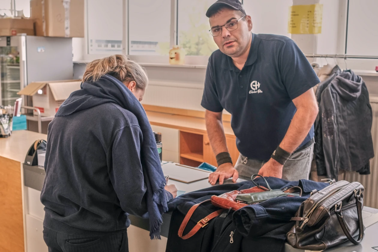 Ein Mann in blauem T-Shirt mit Logo der Erlacher H&ouml;he steht hinter der Theke und blickt direkt zur Kamera, w&auml;hrend eine Frau mit dem R&uuml;cken zur Kamera vor der Theke steht und mit einem Kugelschreiber schreibt. Auf dem Tisch sind Kleidungsst&uuml;cke und eine Handtasche zu sehen.