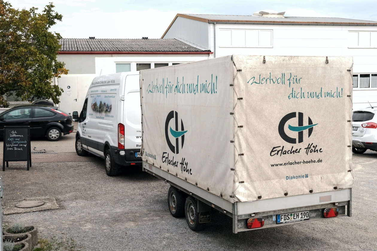 Wei&szlig;er Transporter und Anh&auml;nger mit dem Logo der Erlacher H&ouml;he auf einem Parkplatz vor einem wei&szlig;en Geb&auml;ude.