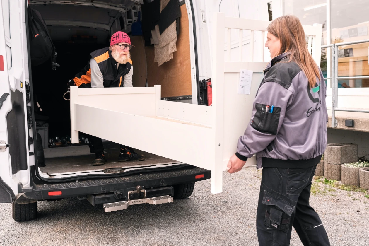 Zwei M&auml;nner laden gemeinsam ein wei&szlig;es M&ouml;belst&uuml;ck aus einem Lieferwagen. Einer der beiden tr&auml;gt eine rote M&uuml;tze, er steht im Fahrzeug, w&auml;hrend der andere au&szlig;erhalb des Fahrzeugs eine graue Jacke mit dem Logo der Erlacher H&ouml;he auf dem R&uuml;cken tr&auml;gt.