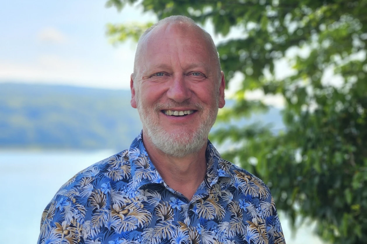 Ein Mann l&auml;chelt freundlich in die Kamera. Er tr&auml;gt ein blaues Hemd mit Blumenmuster. Im Hintergrund sind B&auml;ume und Wasser zu sehen.
