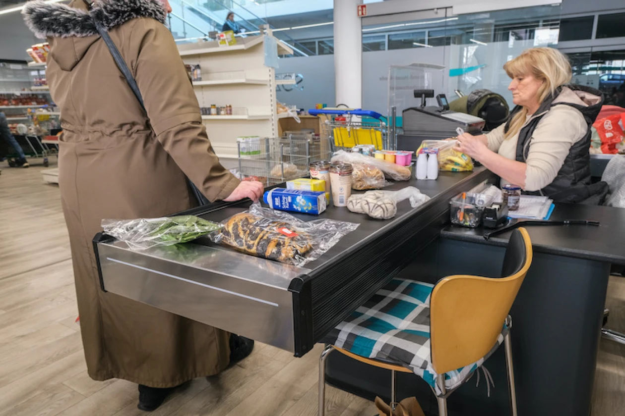 Eine Frau an einer Kasse kassiert den Einkauf einer Kundin ab. Verschiedene Lebensmittel liegen auf dem Kassenband. Eine gelber Stuhl ist im Vordergrund sichtbar.