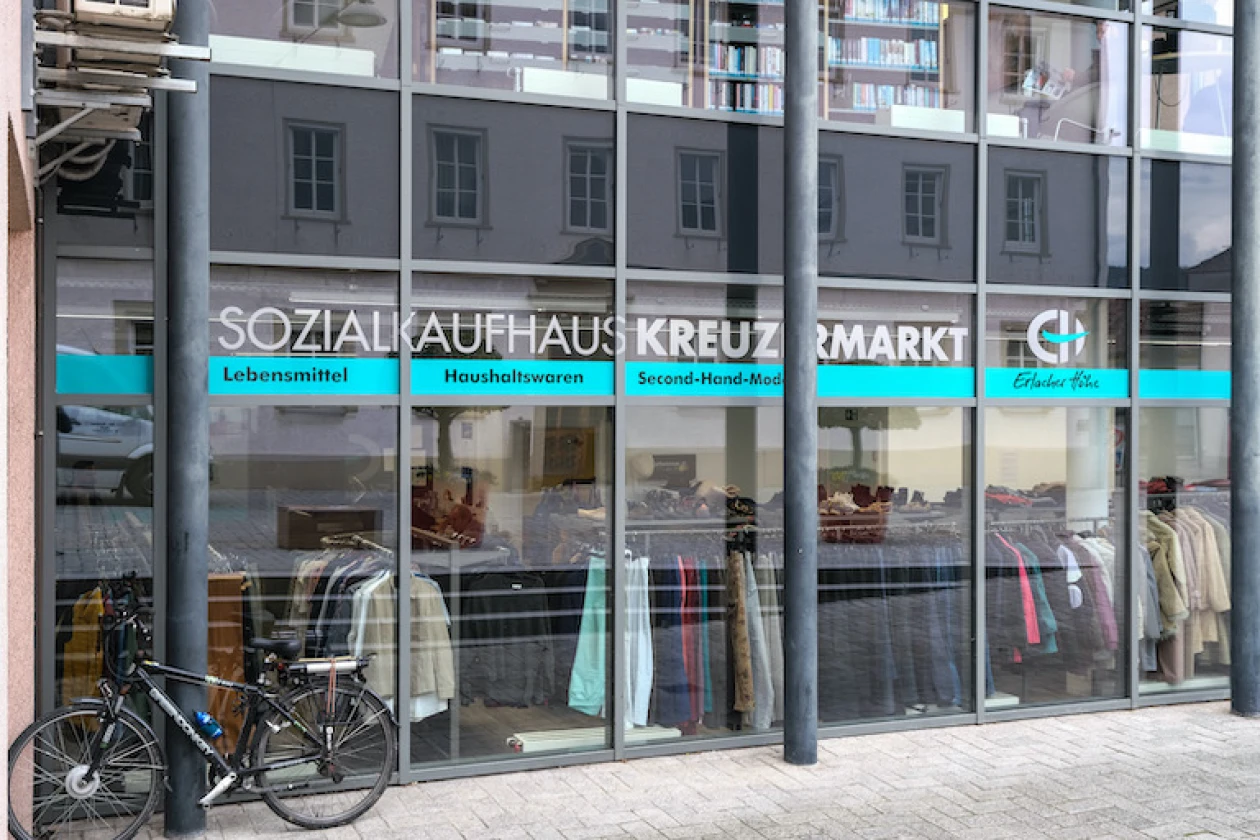 Zu sehen ist die Glasfassade des Sozialaufhauses Kreuzmarkt der Erlacher H&ouml;he mit gro&szlig;em Schriftzug und Logo der Erlacher H&ouml;he im Schaufenster. Kleiderst&auml;nder sind im Innern zu sehen, w&auml;hrend vor dem Laden ein Fahrrad abgestellt ist.