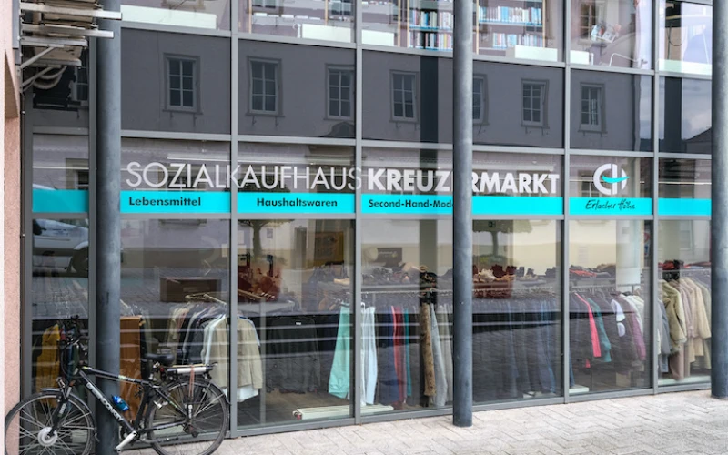 Zu sehen ist die Glasfassade des Sozialaufhauses Kreuzmarkt der Erlacher H&ouml;he mit gro&szlig;em Schriftzug und Logo der Erlacher H&ouml;he im Schaufenster. Kleiderst&auml;nder sind im Innern zu sehen, w&auml;hrend vor dem Laden ein Fahrrad abgestellt ist.