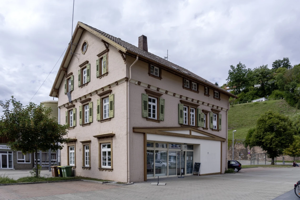 Gro&szlig;es, dreist&ouml;ckiges Geb&auml;ude mit vielen Sprossenfenstern. Das Haus steht zwischen Pflasterfl&auml;chen, B&auml;umen und gepflegter Gr&uuml;nfl&auml;che im Hintergrund. Vor dem Geb&auml;ude ist ein Parkplatz.
