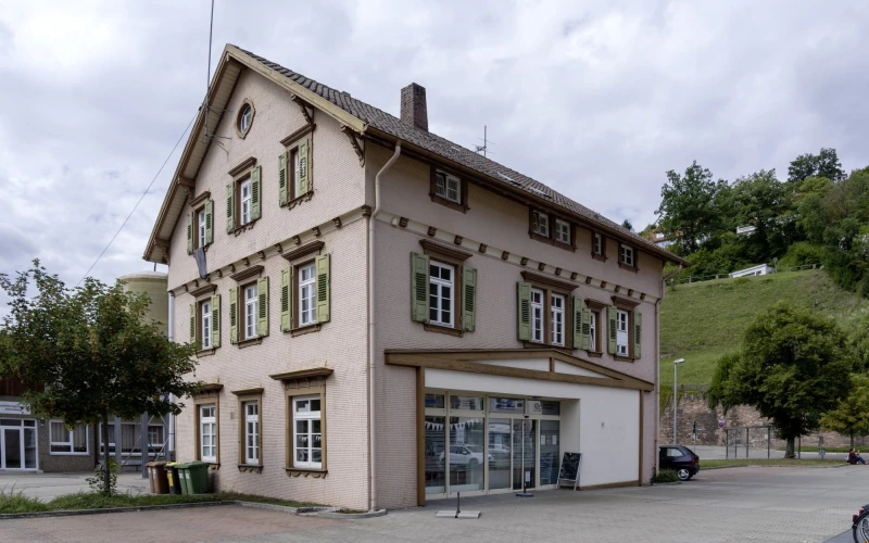 Gro&szlig;es, dreist&ouml;ckiges Geb&auml;ude mit vielen Sprossenfenstern. Das Haus steht zwischen Pflasterfl&auml;chen, B&auml;umen und gepflegter Gr&uuml;nfl&auml;che im Hintergrund. Vor dem Geb&auml;ude ist ein Parkplatz.