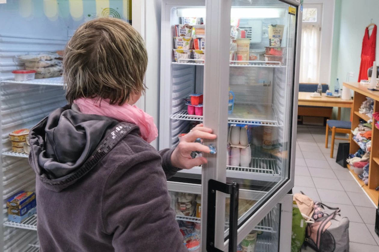 Eine Frau in grauer Jacke steht vor einem offenen K&uuml;hlschrank und blickt auf die verschiedenen Lebensmitteln und Getr&auml;nke. Im Hintergrund sind Holztische, St&uuml;hle und ein Regal zu sehen.