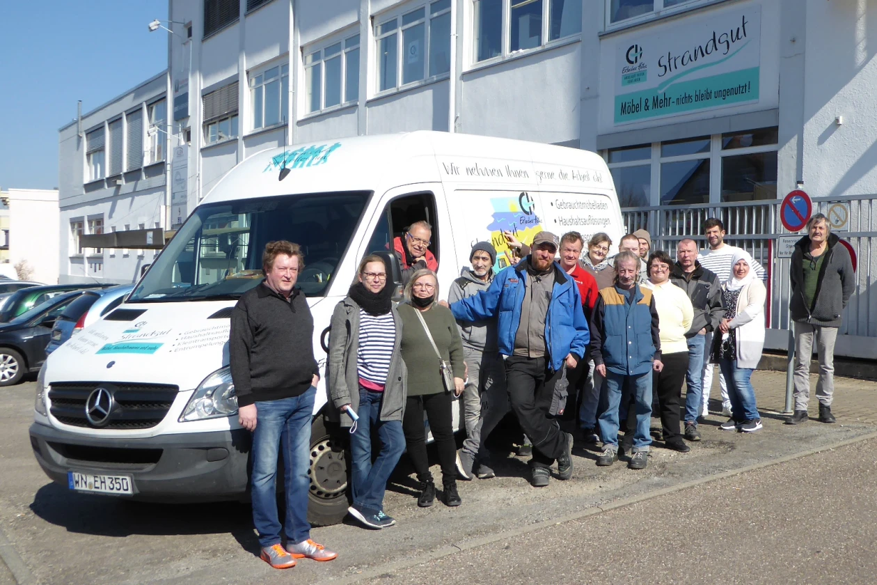 Eine Gruppe von Menschen steht vor einem wei&szlig;en Mercedes-Transporter. Im Hintergrund ist ein Geb&auml;ude mit der Aufschrift "Erlacher H&ouml;he Strandgut" zu sehen.