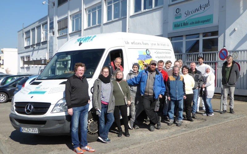 Eine Gruppe von Menschen steht vor einem wei&szlig;en Mercedes-Transporter. Im Hintergrund ist ein Geb&auml;ude mit der Aufschrift "Erlacher H&ouml;he Strandgut" zu sehen.