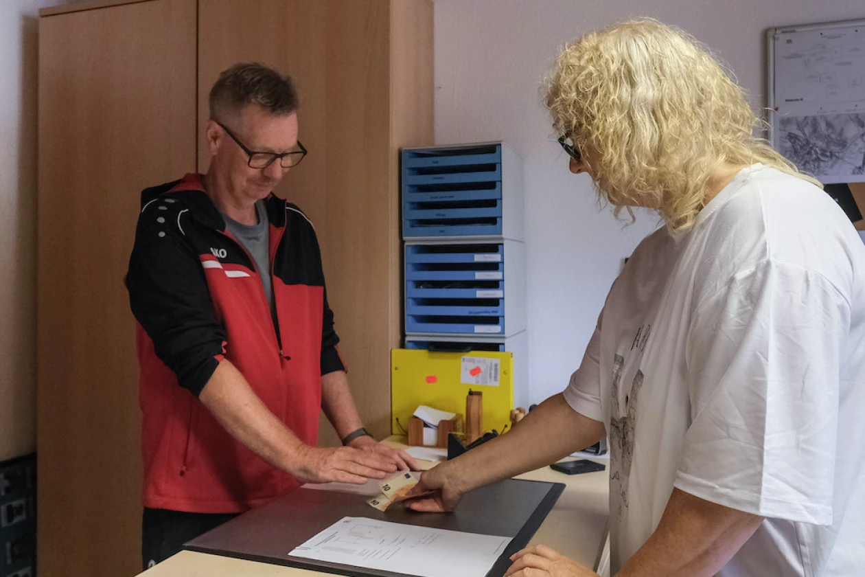 Ein Mann in einer rot-schwarzen Jacke und eine Frau in einem wei&szlig;en T-Shirt stehen an einem Schreibtisch. Die Frau &uuml;bergibt dem Mann zwei Zehn-Euro-Scheine. Im Hintergrund sind ein Holzschrank und eine blaue Ablage zu sehen.